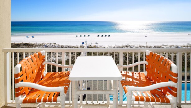 A balcony with a white table and orange wicker chairs, overlooking a beach with a clear blue sky and a swimming pool. - Waters Edge 514 • Beachfront Balcony, Platinum Condo, Covered Prkg, Free Beach Service (Fort Walton Beach)