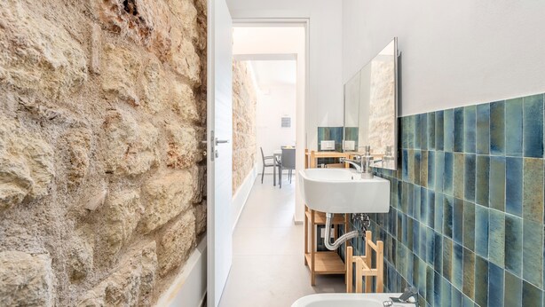 Un baño estrecho con pared de piedra, lavamanos blanco y pared revestida de azulejos azules.