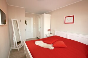 Une chambre à coucher avec une couvre-lit rouge, des meubles blancs, une horloge au mur et un miroir.