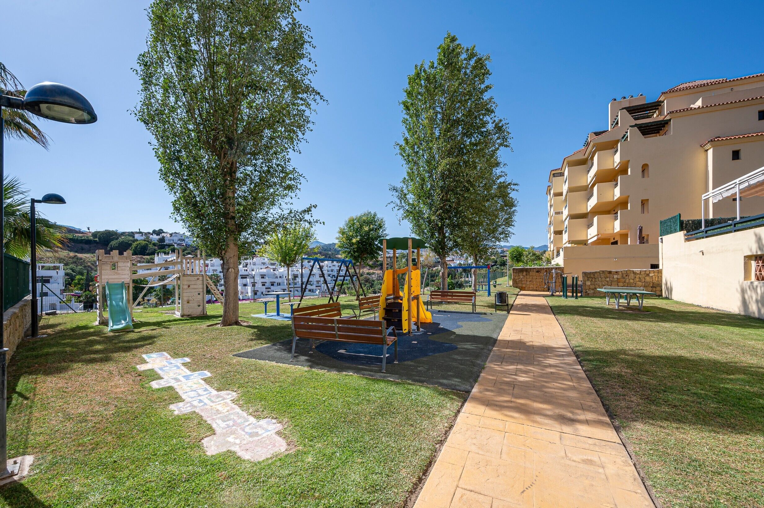 A playground with swings and a slide, a paved path, and a building with balconies.