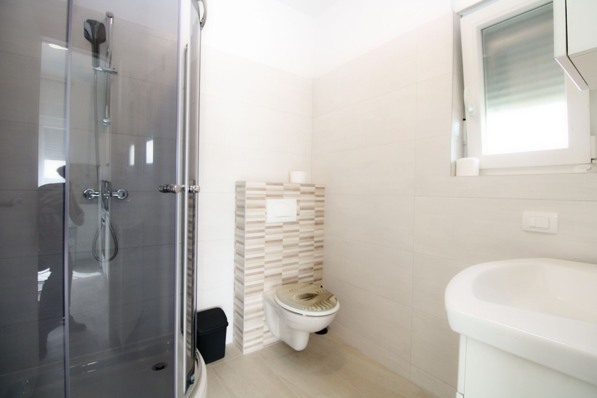 A modern bathroom with a glass shower enclosure, a toilet with a beige patterned tile wall, and a white sink.