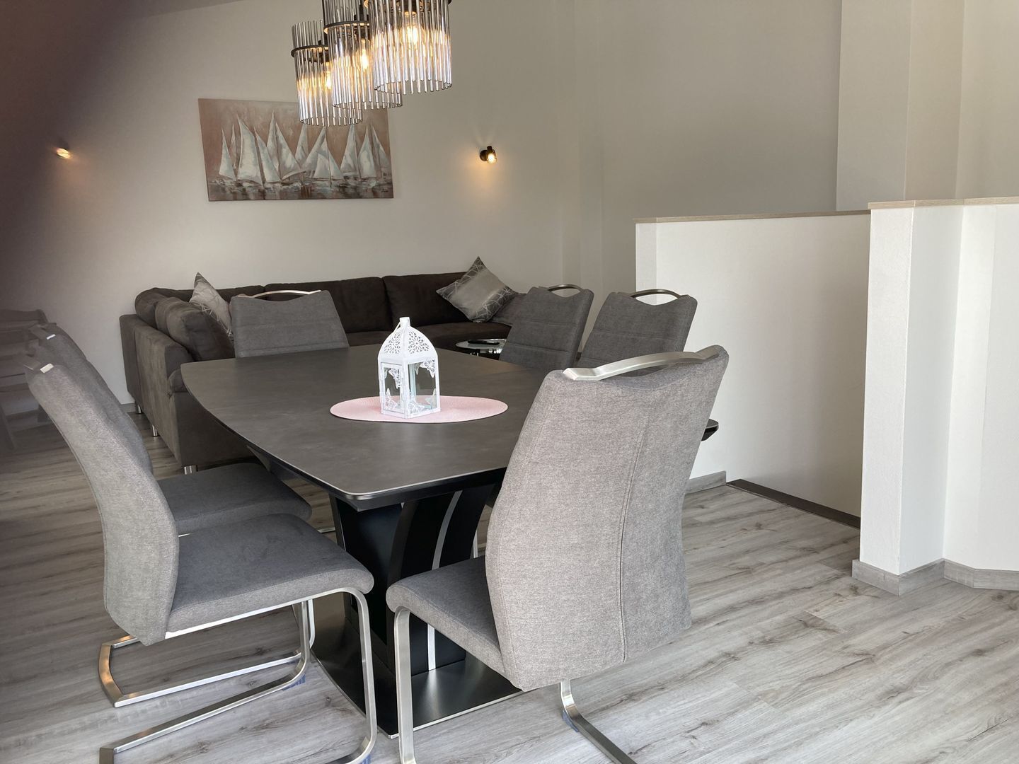 A modern dining area with a rectangular table, six grey upholstered chairs, a white decorative lantern, and a chandelier.