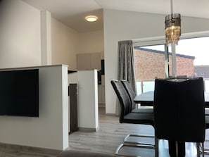 Una cocina moderna con una televisión de pantalla plana, una zona de comedor con mesa y sillas, y un amplio ventanal con vistas a un edificio de ladrillo.