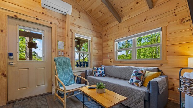 Interior acolhedor de uma cabana, com fachada de madeira, um sofá, uma poltrona, uma mesa pequena e uma janela com vista para as árvores.