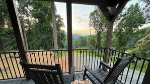 Um deck de madeira com duas cadeiras e uma mesa pequena, com vista para uma paisagem cênica com árvores e uma estrada.