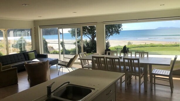 A modern kitchen with a large island, dining area with white chairs, and a view of the beach. - Absolute beachfront! Sleeps 12+
Built 2011 (Waihi Beach)