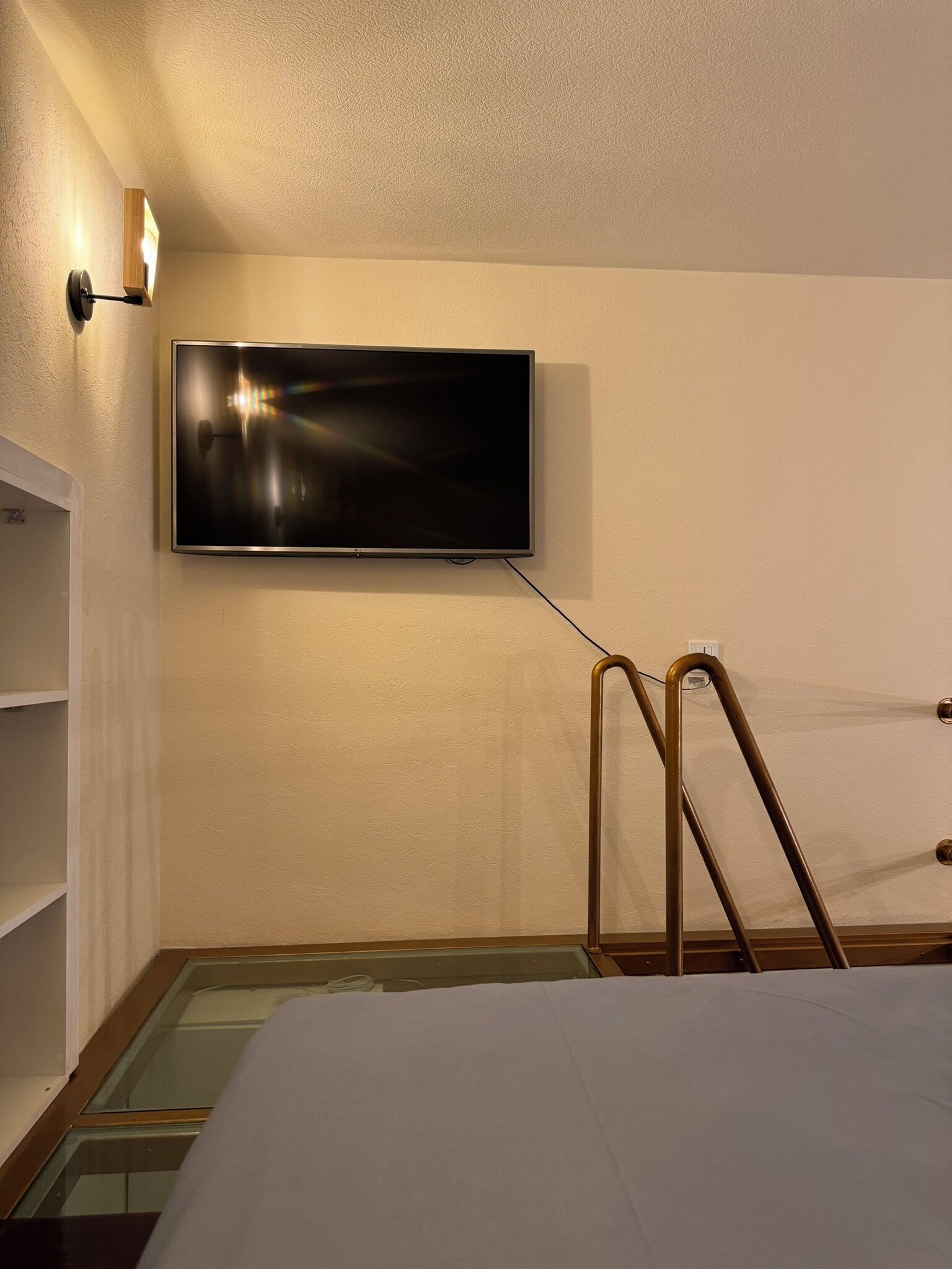 A hotel room with a flat-screen TV mounted on the wall, a wooden ladder, and a glass floor.