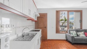 A kitchen with white cabinets, a built-in stove, and a sink. There is a wooden door and a window with a view of palm trees. - Vacation Apartment 'Apto Veracruz' with Shared Pool, Private Terrace and Wi-Fi (Costa Adeje)