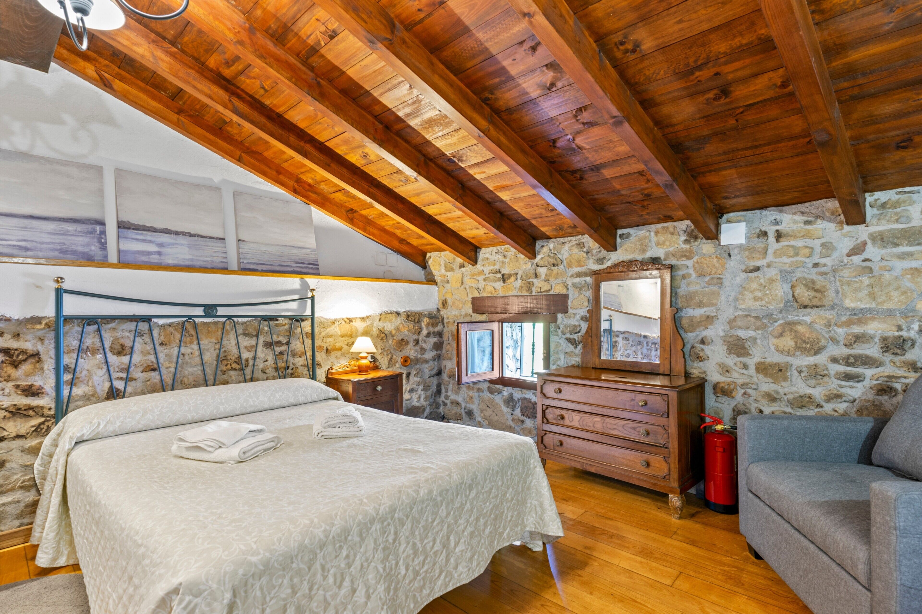 Un dormitorio con techo de madera, paredes de piedra, una cama, un aparador, un espejo y un extintor.