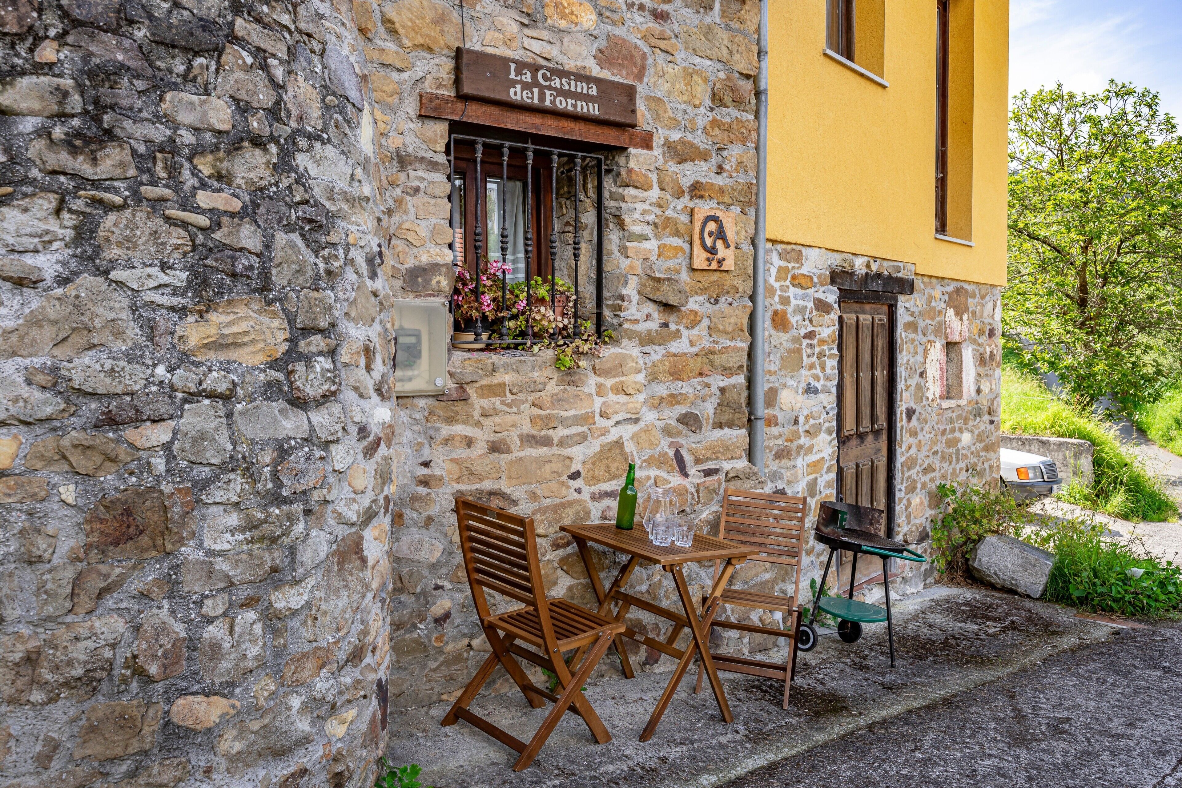 A stone building with a wooden table and chairs, a window with flowers, and a sign reading 'La Casina del Fornu.'