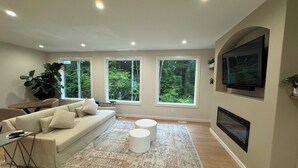 Una sala de estar moderna con un sofá, una mesa de centro y una chimenea. Hay ventanas grandes con vistas a la vegetación.