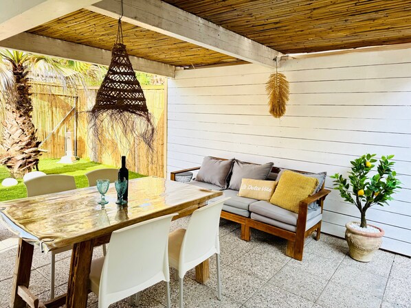 A patio with a wooden table, white chairs, a sofa with cushions, a hanging light fixture, and a potted plant.