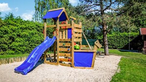 Une structure de jeux en bois, avec un toboggan bleu et une zone d’escalade, installée sur un sol sablonneux, avec des arbres en arrière-plan.