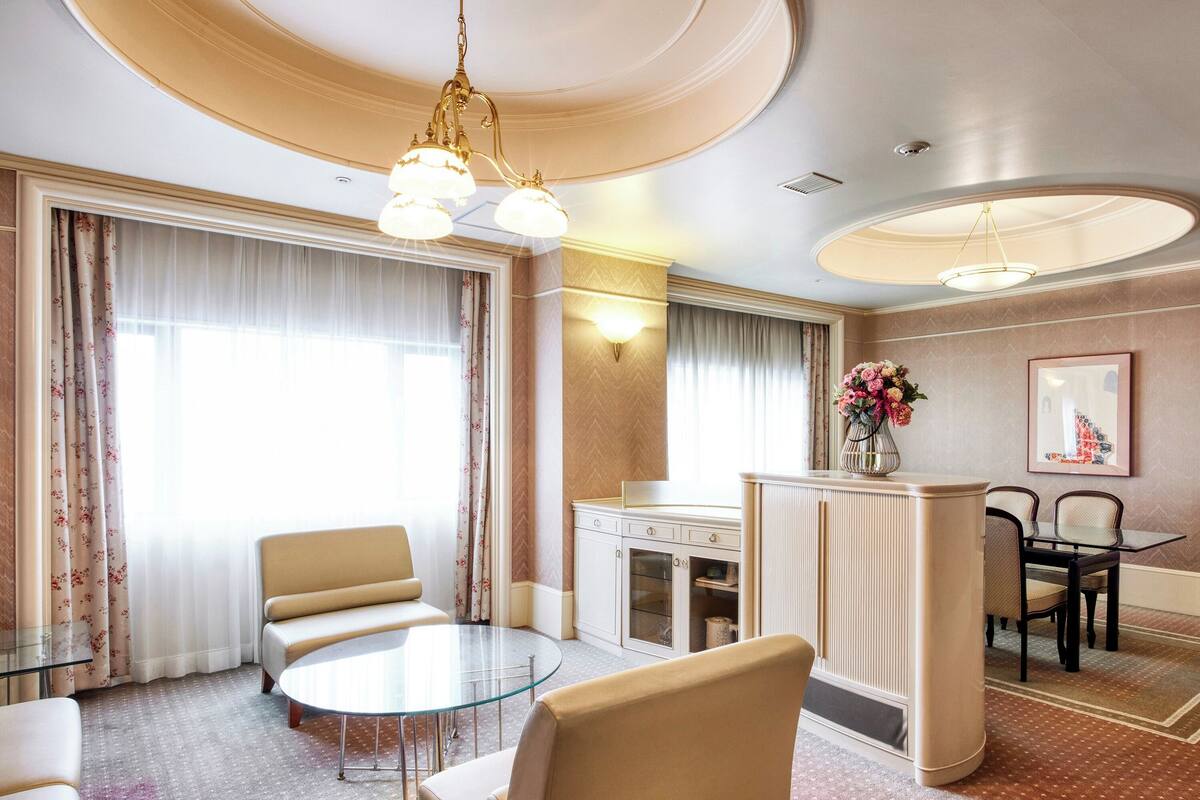 A hotel room with a round glass table, beige chairs, a minibar, a dining area with a table and chairs, and a large window with curtains.