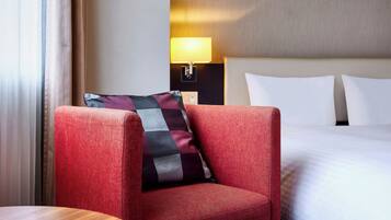 A hotel room with a red armchair, a round wooden table, a bed with white linens, and a nightstand with a lamp.