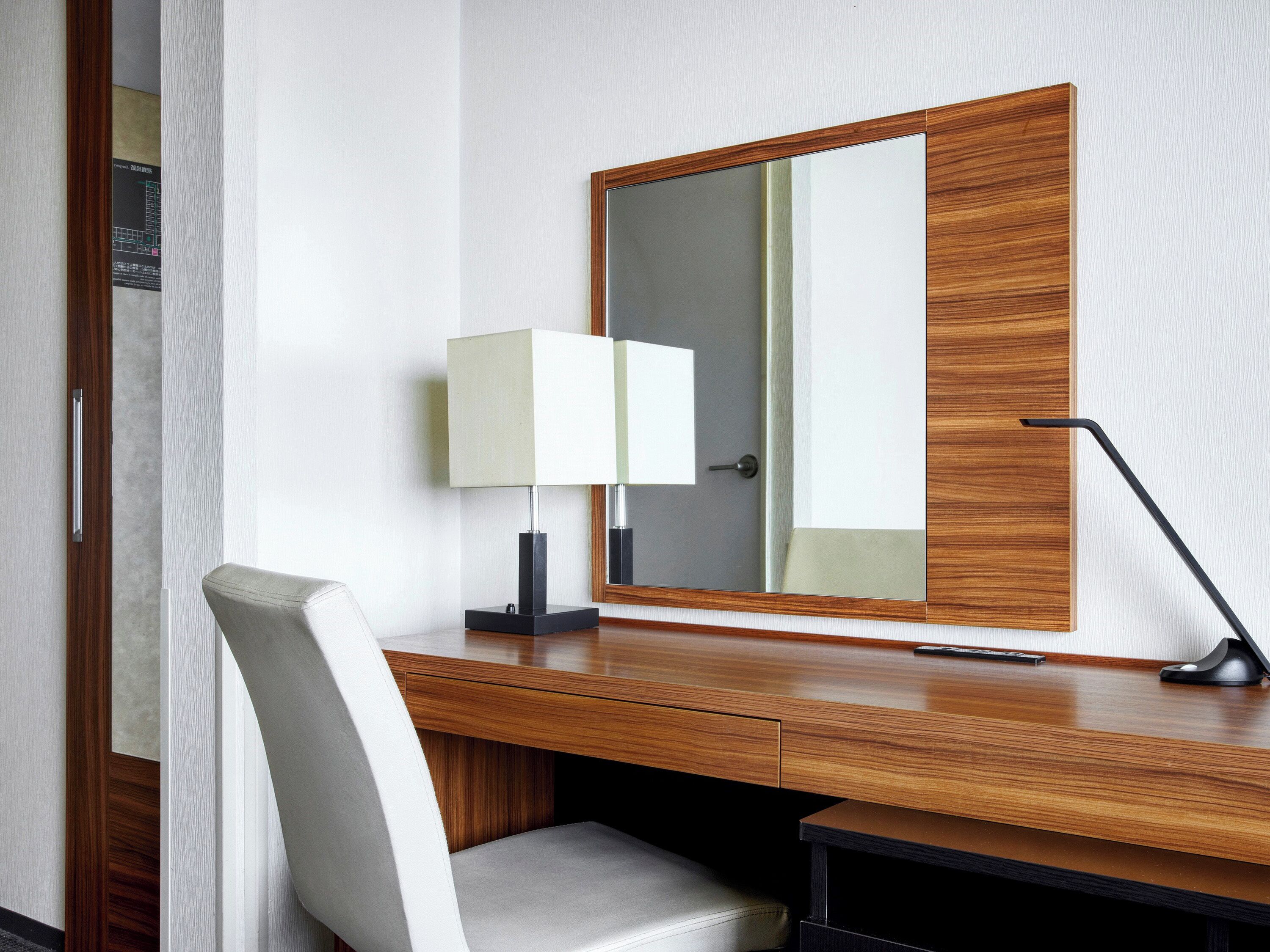 A hotel room with a wooden desk, a white chair, a lamp, and a mirror.
