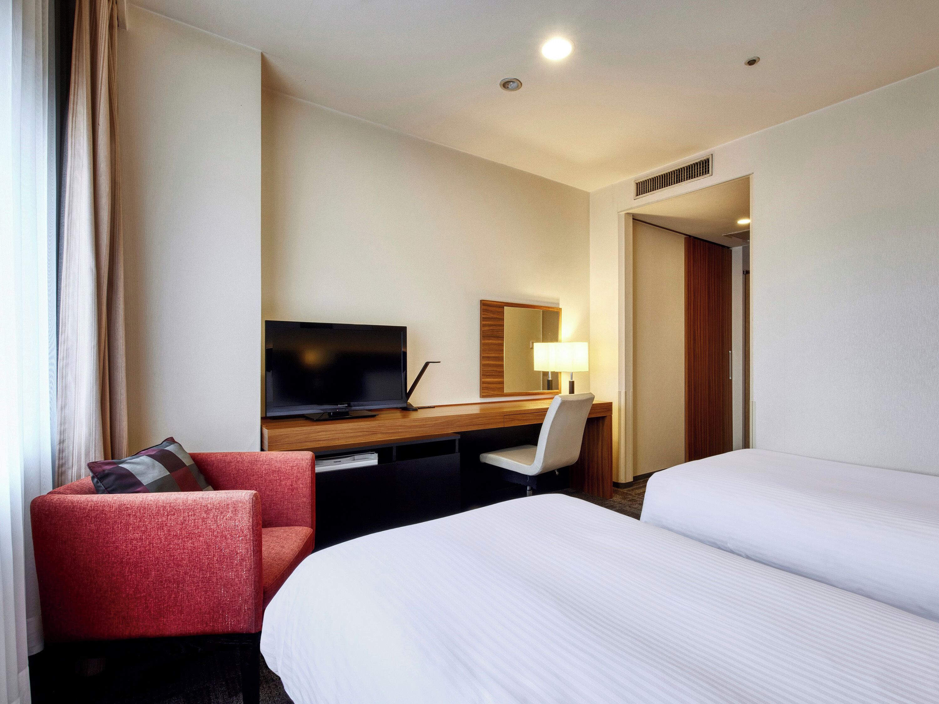 A hotel room with a bed, a desk with a television, a chair, and a bedside table with a lamp.