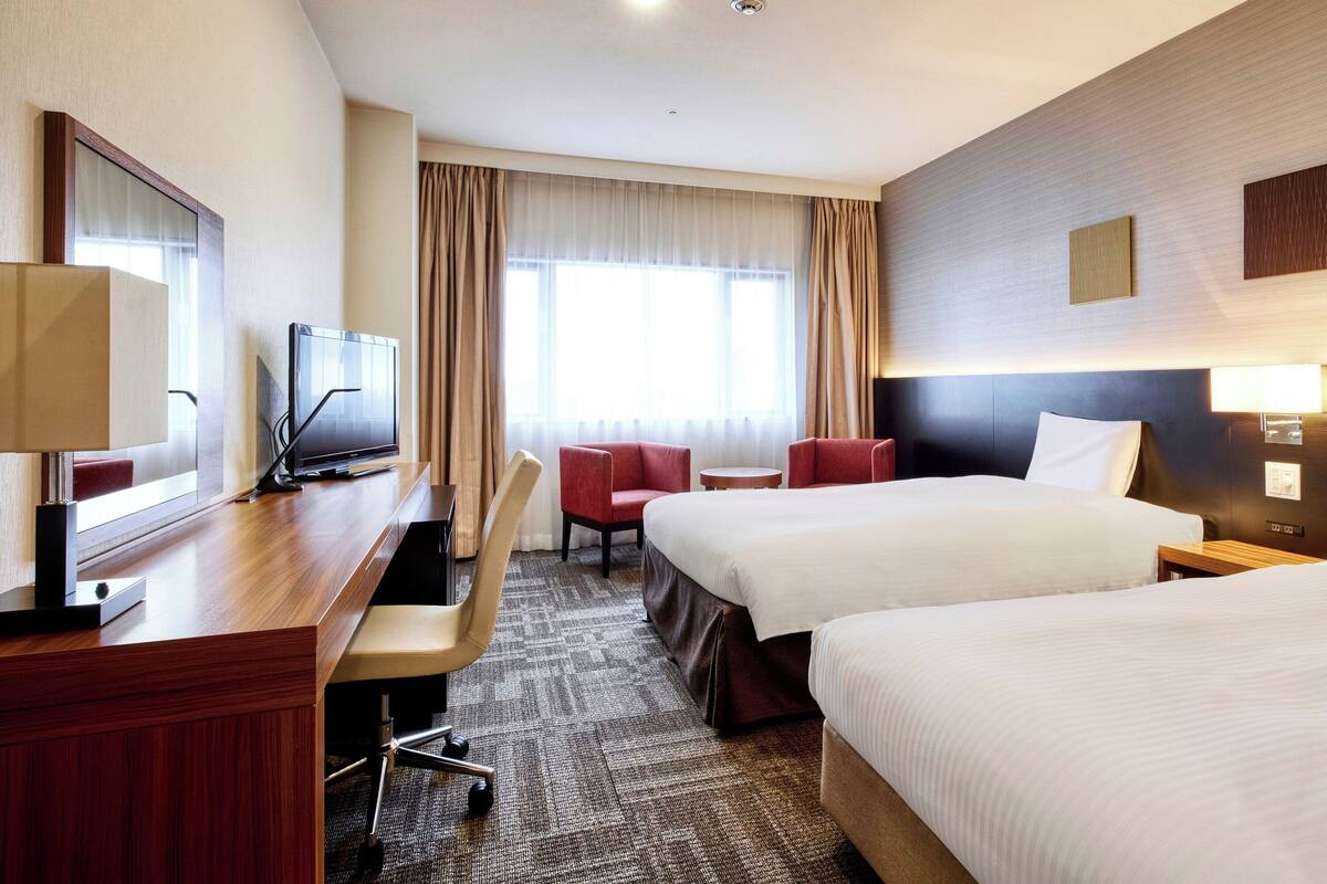 A hotel room with two beds, a desk with a TV, a chair, and a small table.