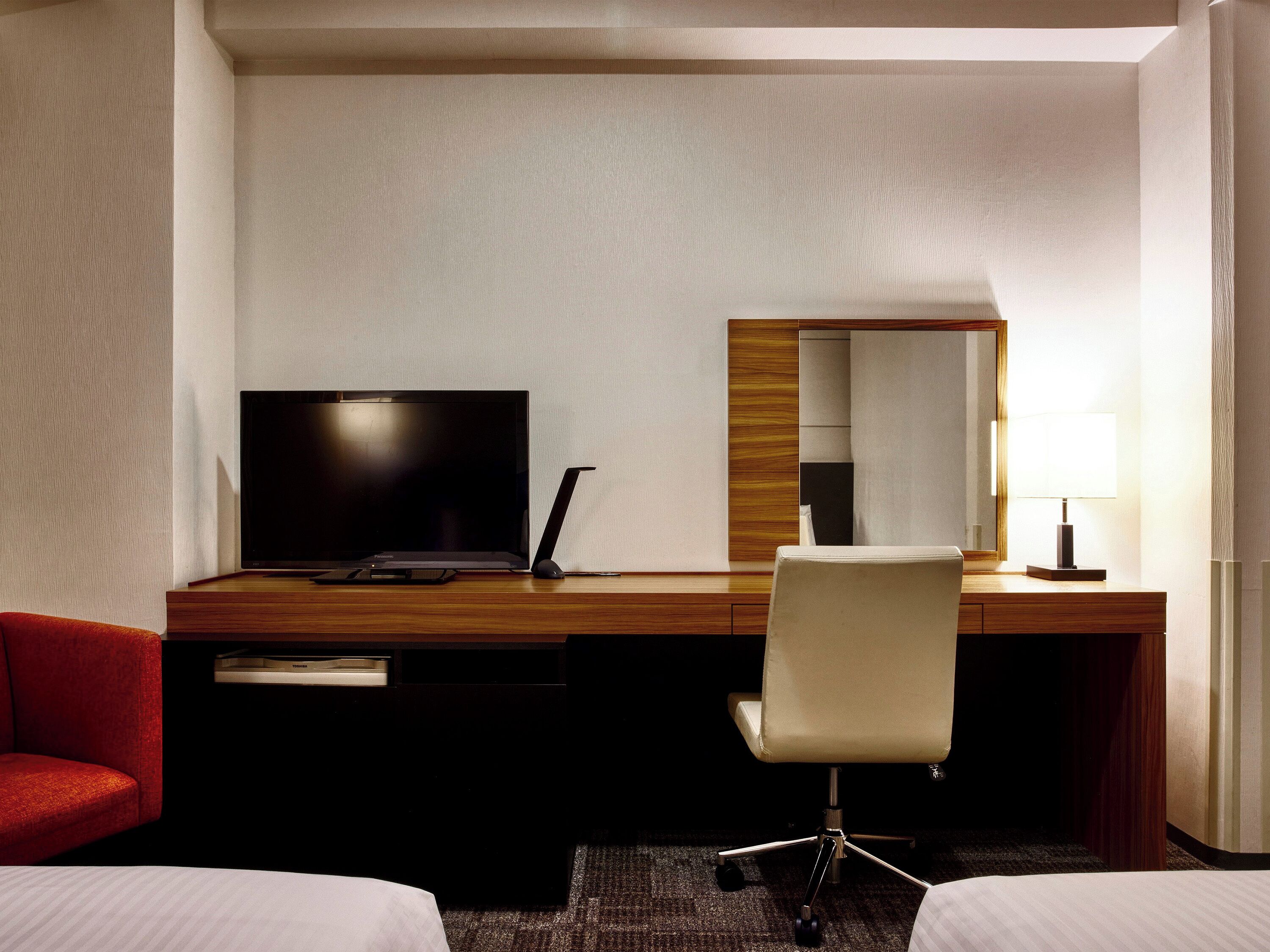 A hotel room with a wooden desk, a television, a chair, a mirror, and a lamp.