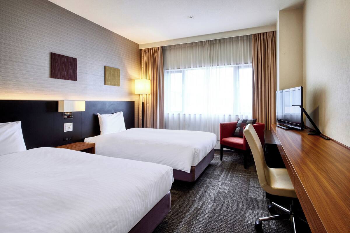 A hotel room with two beds, a desk, a chair, a TV, and a window with curtains.