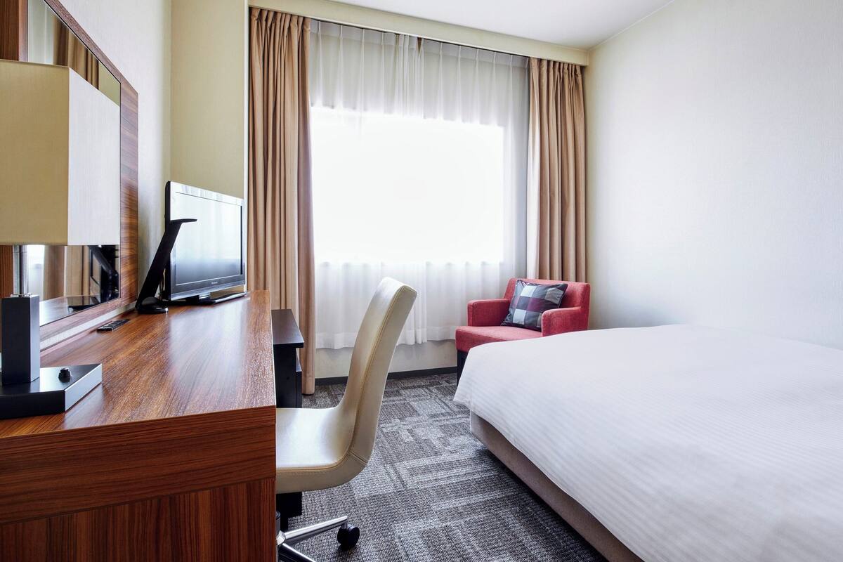 A hotel room with a bed, a desk with a TV, a chair, and a window with curtains.