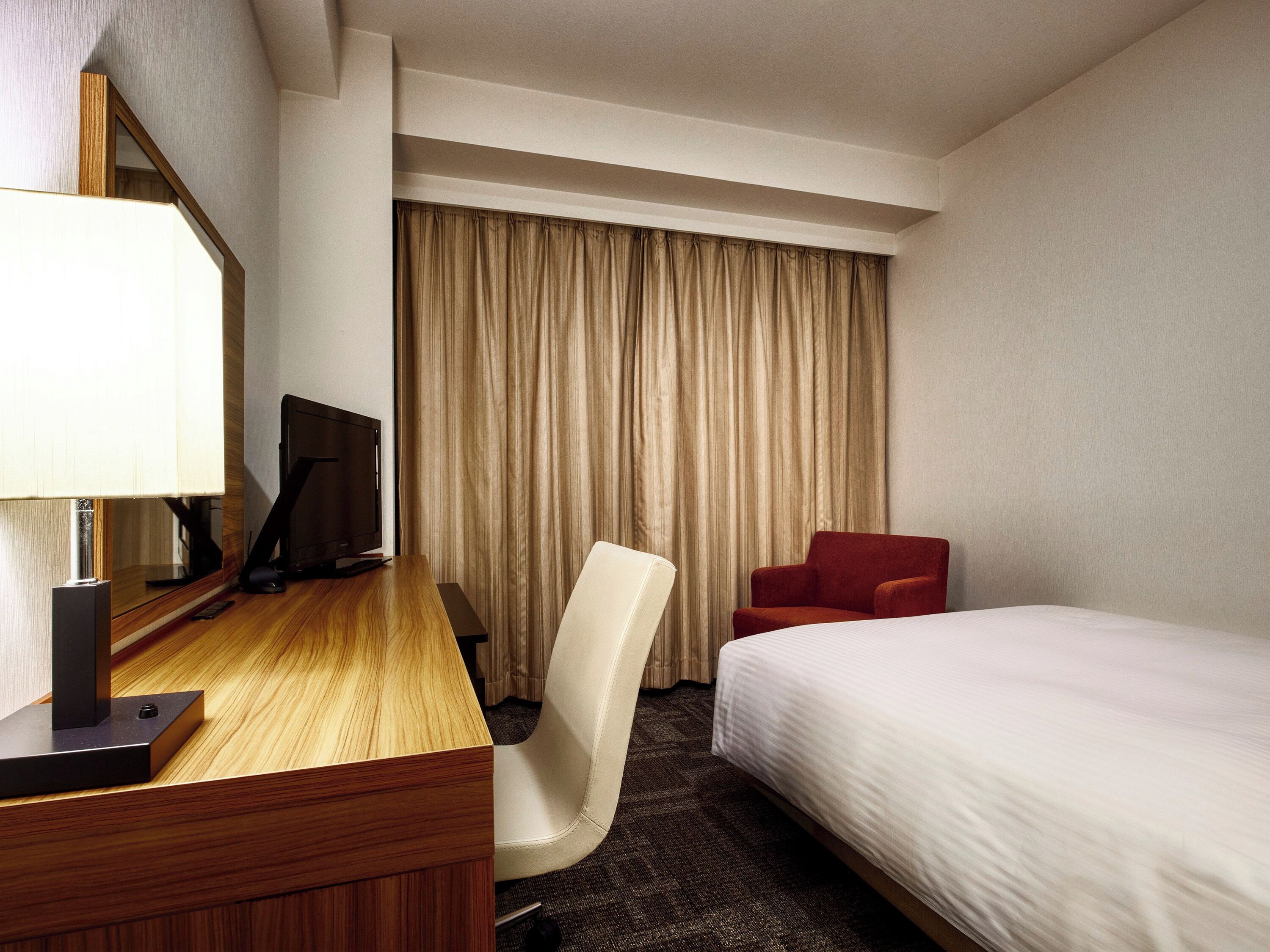 A hotel room with a wooden desk, a white chair, a red chair, a bed, a TV, and a window with curtains.
