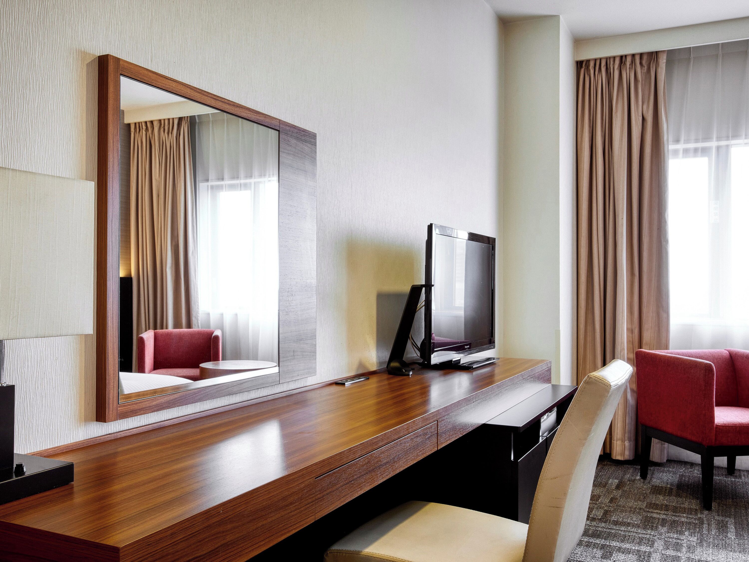 A hotel room with a wooden desk, a television, a chair, and a large mirror.