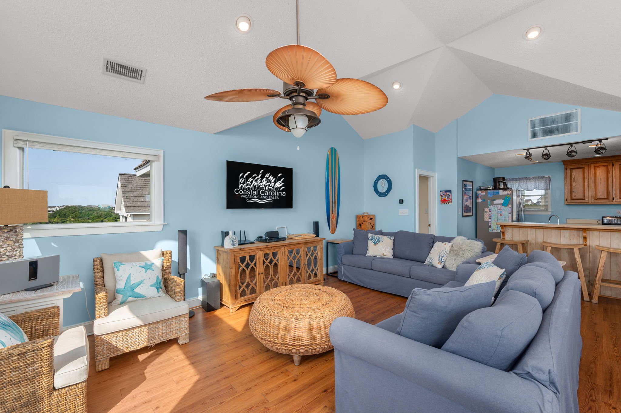 A spacious living area with a large ceiling fan, a grey sofa, wicker furniture, and a kitchen area with wooden cabinets.