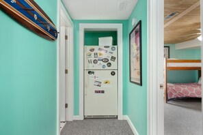 A narrow hallway with a white refrigerator covered in stickers, a bunk bed in the background, and a framed picture on the wall. - Wanderer's Rest: Oceanside, Private Pool, Hot Tub, Close to Beach! (Nags Head)