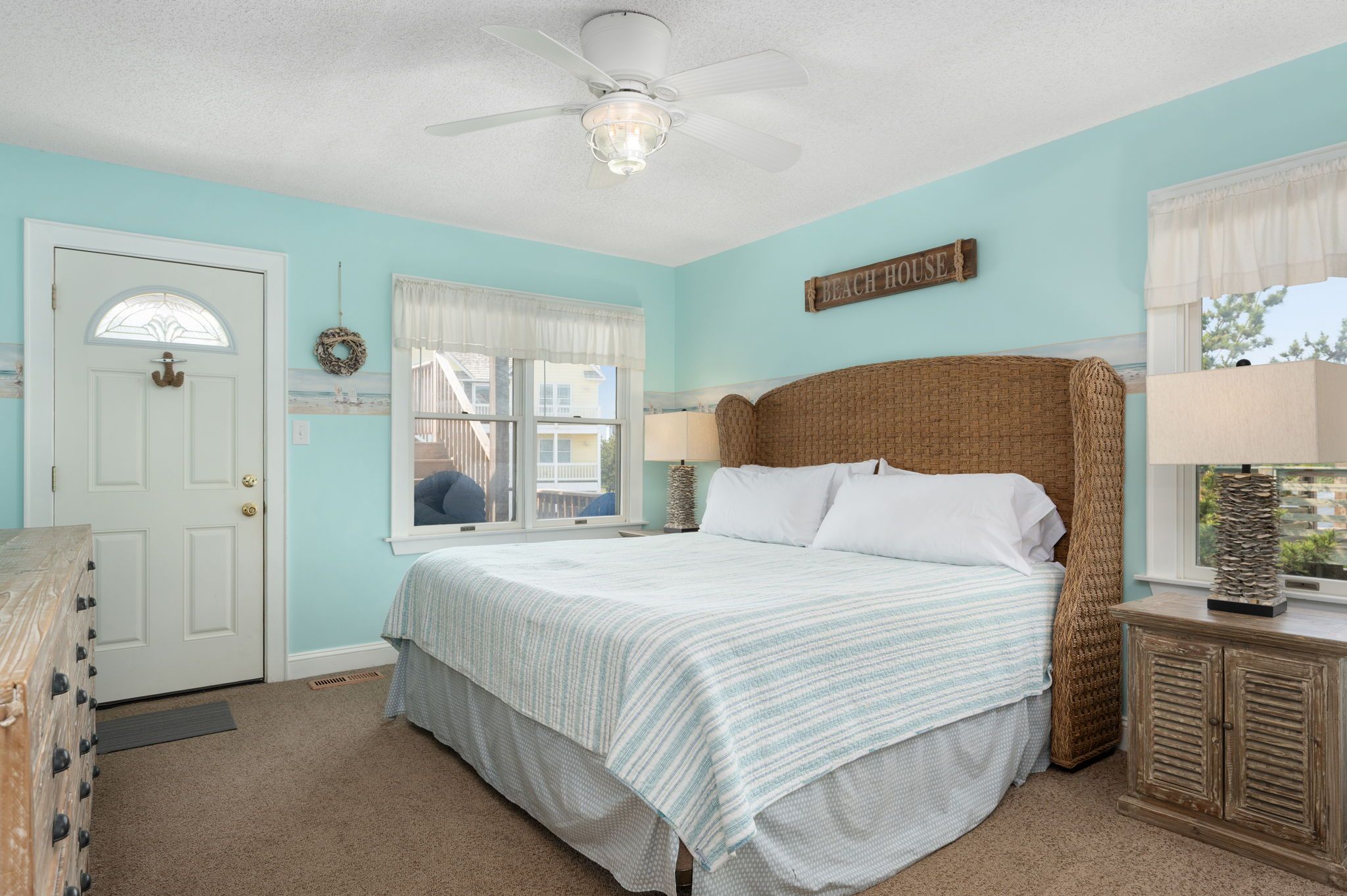 A bedroom with a bed, a ceiling fan, a window with curtains, a door, and a wall sign that reads "BEACH HOUSE".