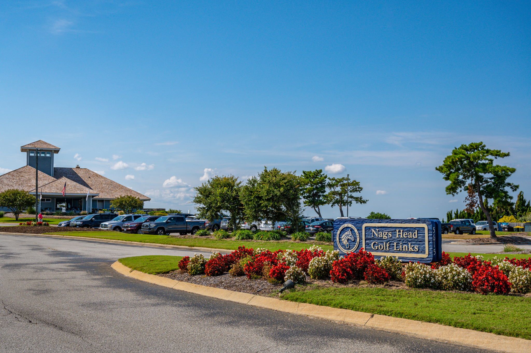 A golf course sign with "Nags Head Golf Links" written on it, surrounded by a landscaped area with red and white flowers.