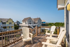 A wooden deck with white Adirondack chairs, a wooden railing, and a view of houses in the background. - Wanderer's Rest: Oceanside, Private Pool, Hot Tub, Close to Beach! (Nags Head)