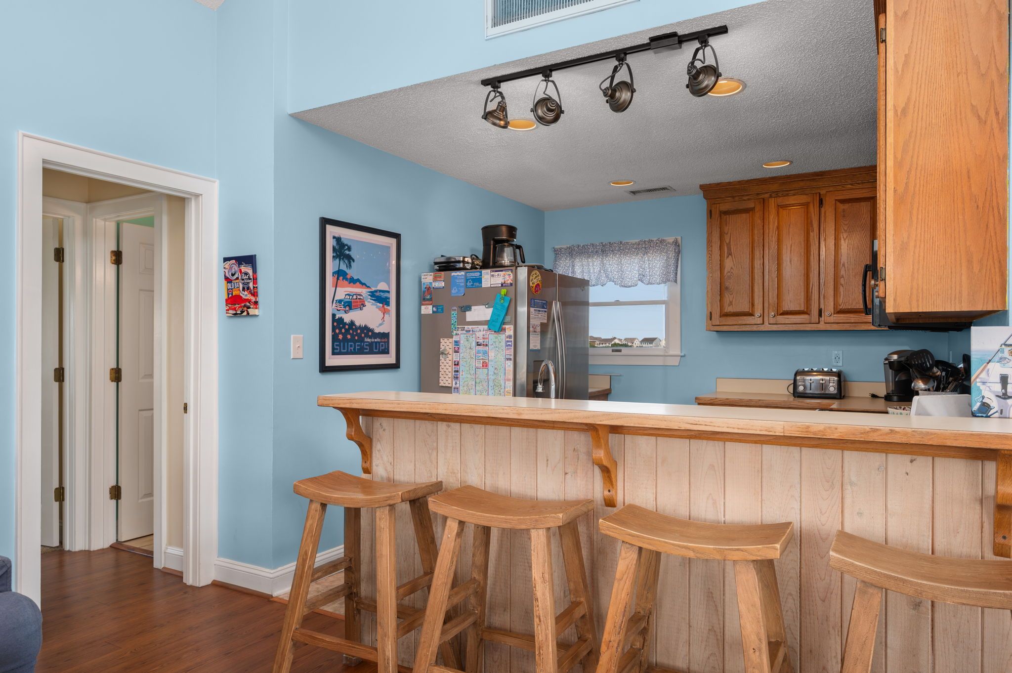 A kitchen with wooden bar stools, a refrigerator, and a window with a lace curtain.