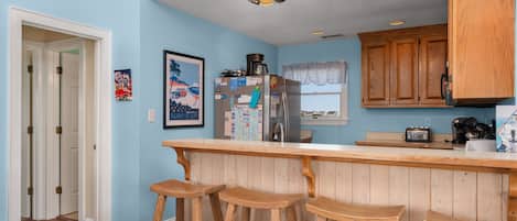 A kitchen with wooden bar stools, a refrigerator, and a window with a lace curtain.