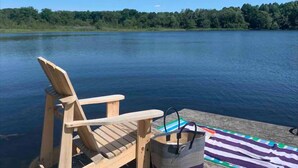 Une chaise en bois et un sac à rayures sont posés sur un quai, au bord d’un lac paisible, avec une forêt en arrière-plan.