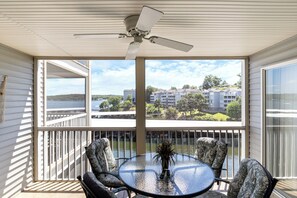 A balcony with a glass table and chairs, a ceiling fan, and a view of a lake and buildings. - Games, Lake Views, Ease of Access to Unit & Attractions, Family-Friendly, Outdoor Comfort (Lake Ozark)