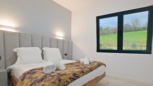 A modern bedroom with a bed, bedside tables, a window with a view of a green field, and a headboard with built-in lighting.