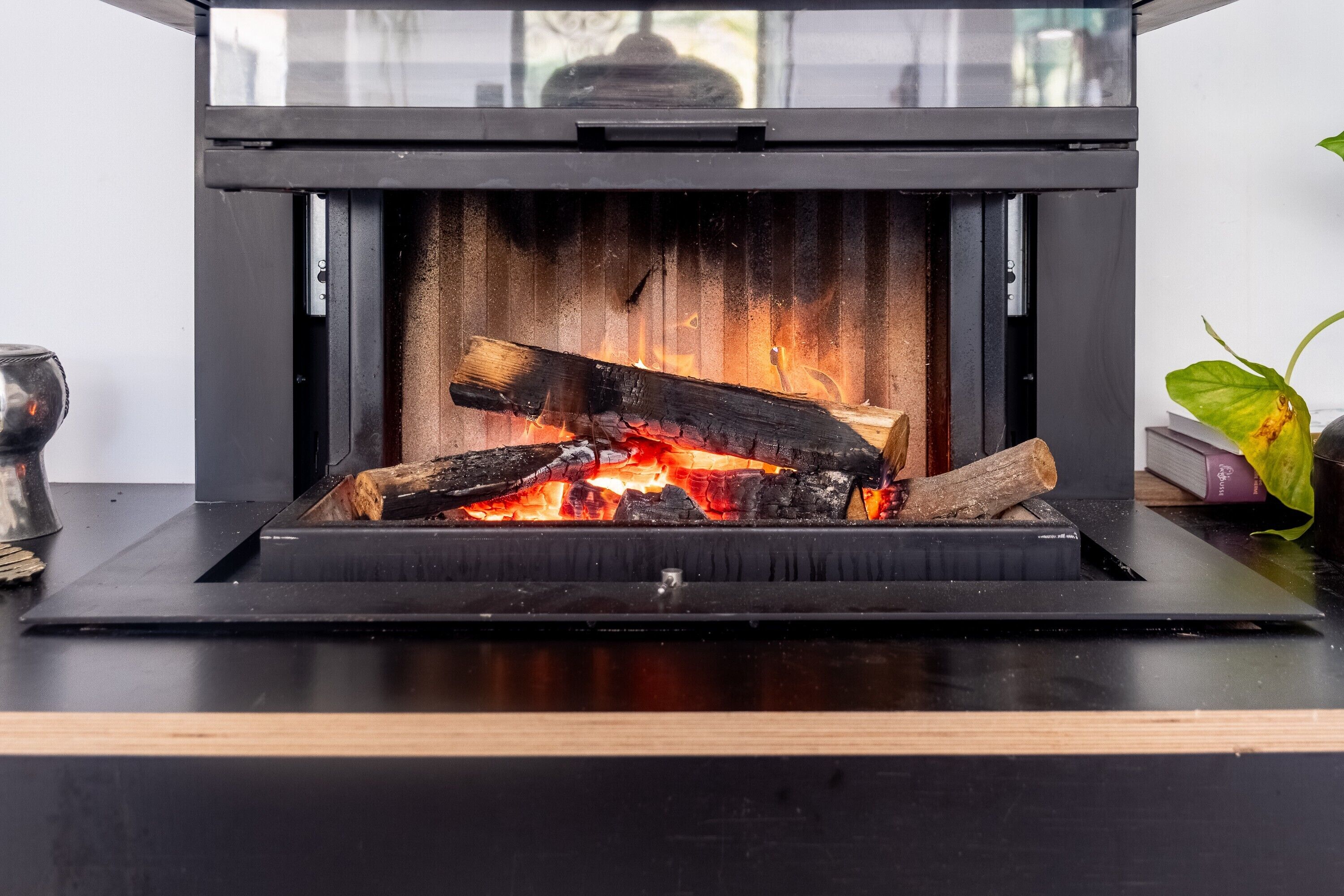A modern fireplace with a glass enclosure, a wooden mantel, and a visible fire burning logs.