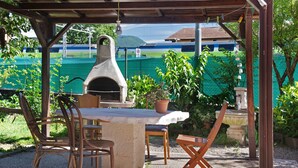Un patio al aire libre con una mesa de piedra, sillas, un horno de pizza y vistas a un campo verde y un tren al fondo.