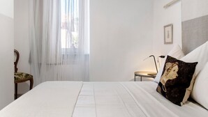 Una cama bien hecha con ropa blanca, una silla con un cojín estampado, una mesita de noche con una lámpara y una cortina translúcida sobre una ventana.