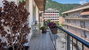 Un balcon agrémenté de plantes en pot, avec une table et des chaises, donnant sur une rue bordée d’immeubles et surmontée, au loin, d’une montagne.