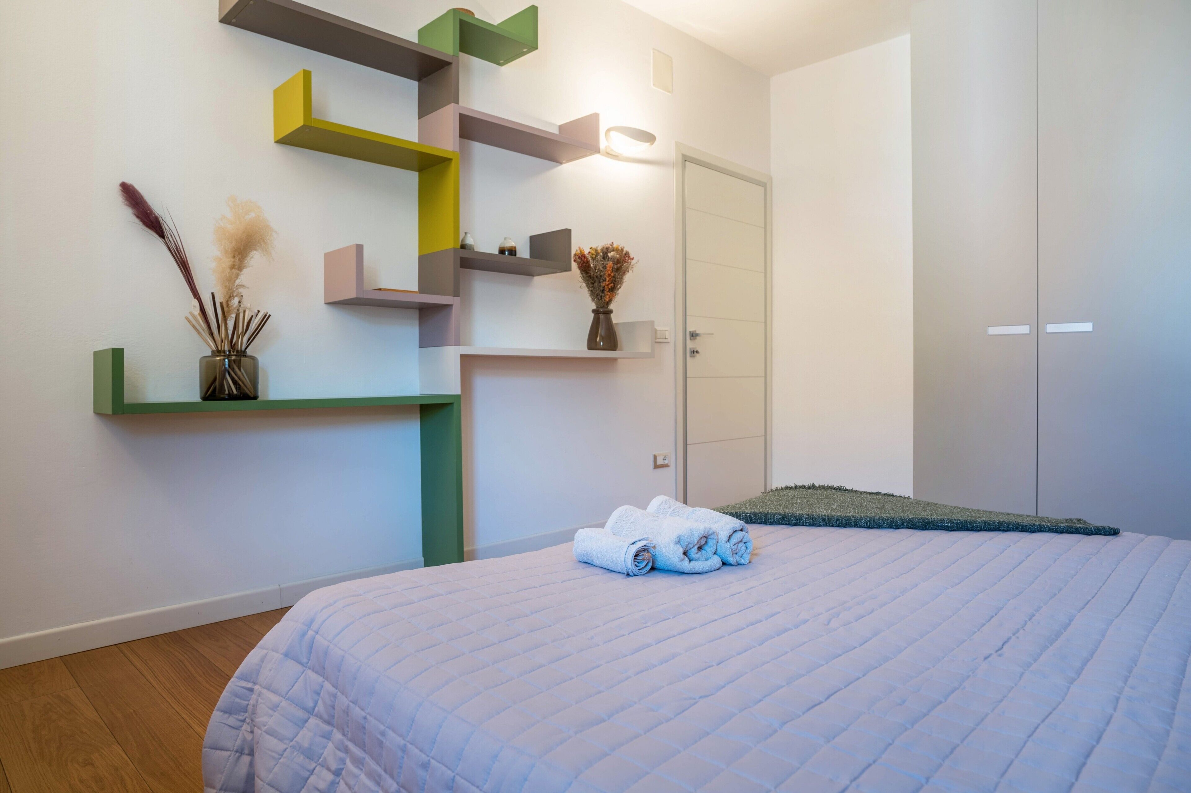 Un dormitorio con cama, estante con objetos decorativos, una puerta y una alfombra pequeña.