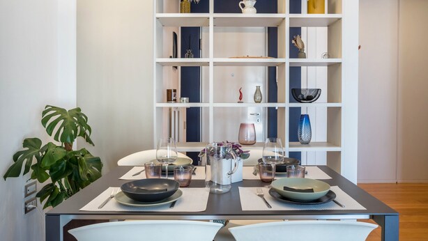 Un coin repas avec une table dressée pour deux personnes, une bibliothèque agrémentée de divers objets décoratifs et une plante en pot.