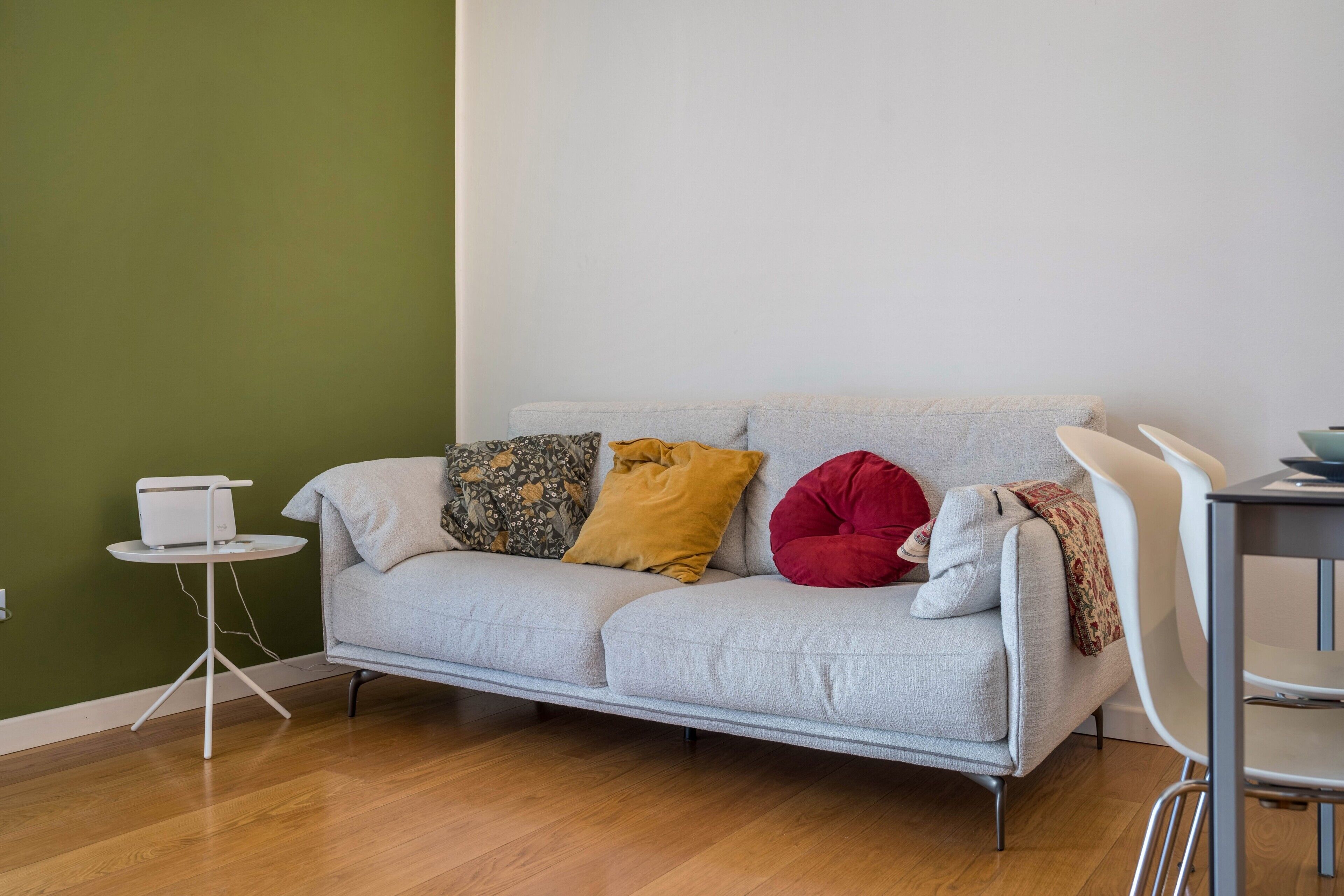 Een woonkamer met een grijze bank, een klein rond tafeltje met een wit apparaat, en een witte stoel.