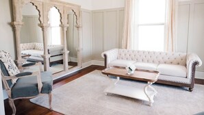 Une pièce comprenant un canapé blanc recouvert de tissu, une table basse en bois, un grand miroir avec un cadre orné, une chaise en bois et un lustre.