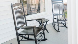 Une véranda avec deux fauteuils à bascule et une petite table, un revêtement extérieur blanc et une fenêtre ornée d’un treillis décoratif.