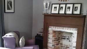 A bedroom with a brick fireplace, a purple armchair, a white bedspread, and a mirror with a sunburst frame. - 1874 House. Walk Everywhere. Pets Welcome. (Beaufort)