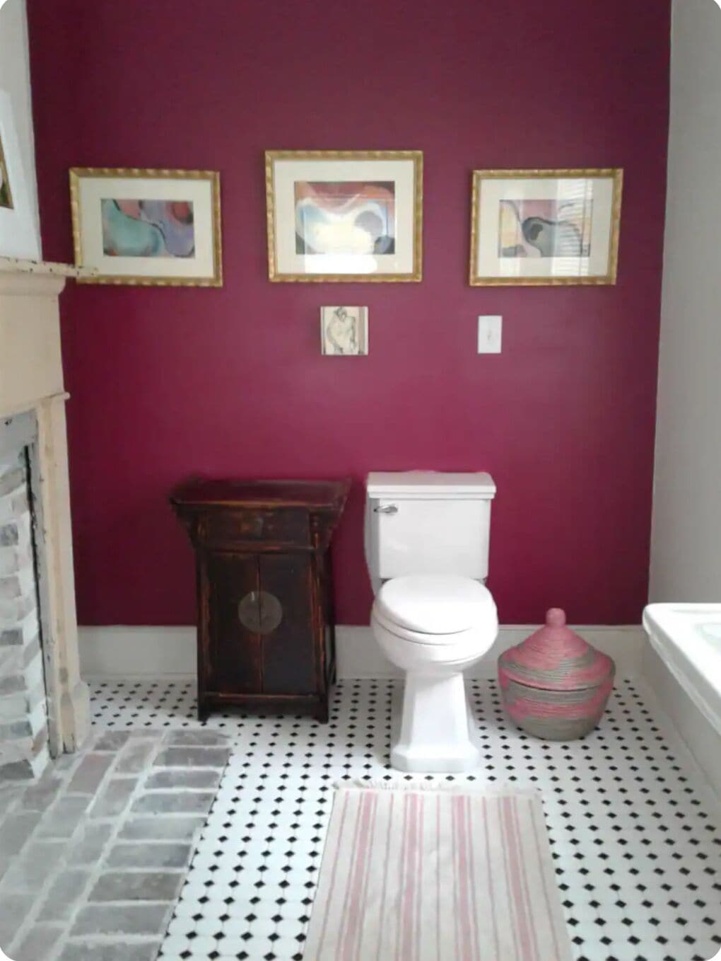 A bathroom with a white toilet, a wooden cabinet, and a pink trash bin. There are three framed pictures on the wall.