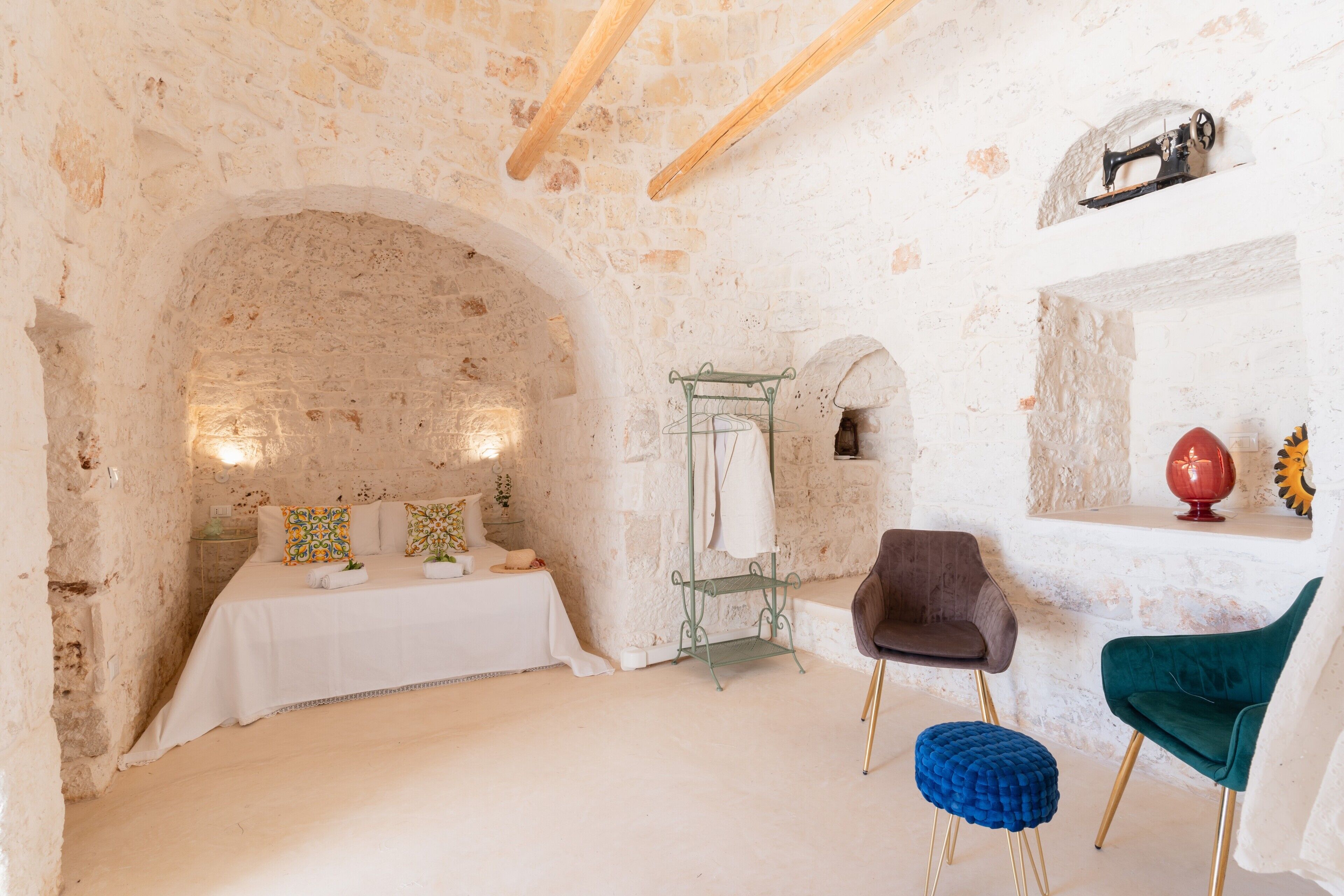 A stone-walled room with a bed, a chair, a small stool, and a shelf with decorative items.