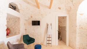 A room with exposed stone walls, a television mounted on the wall, a green chair, a grey chair, and a small stool.